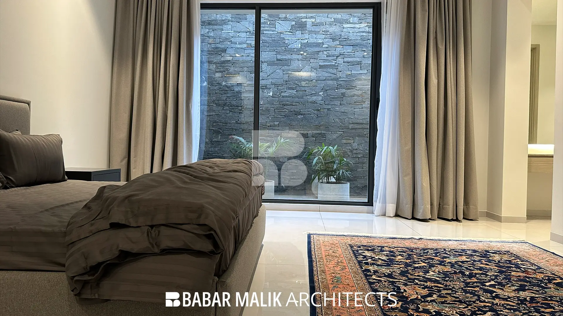 Bedroom interior with a neutral color palette, featuring a large glass sliding door that opens to a private internal courtyard clad in dark, textured stone.