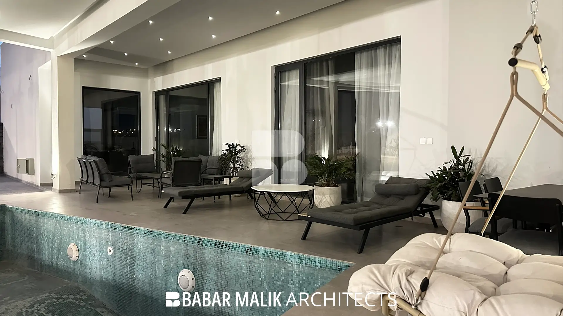 A covered outdoor poolside patio space featuring dark grey sun loungers, outdoor seating, a modern hanging chair, and a mosaic-tiled swimming pool in the foreground.