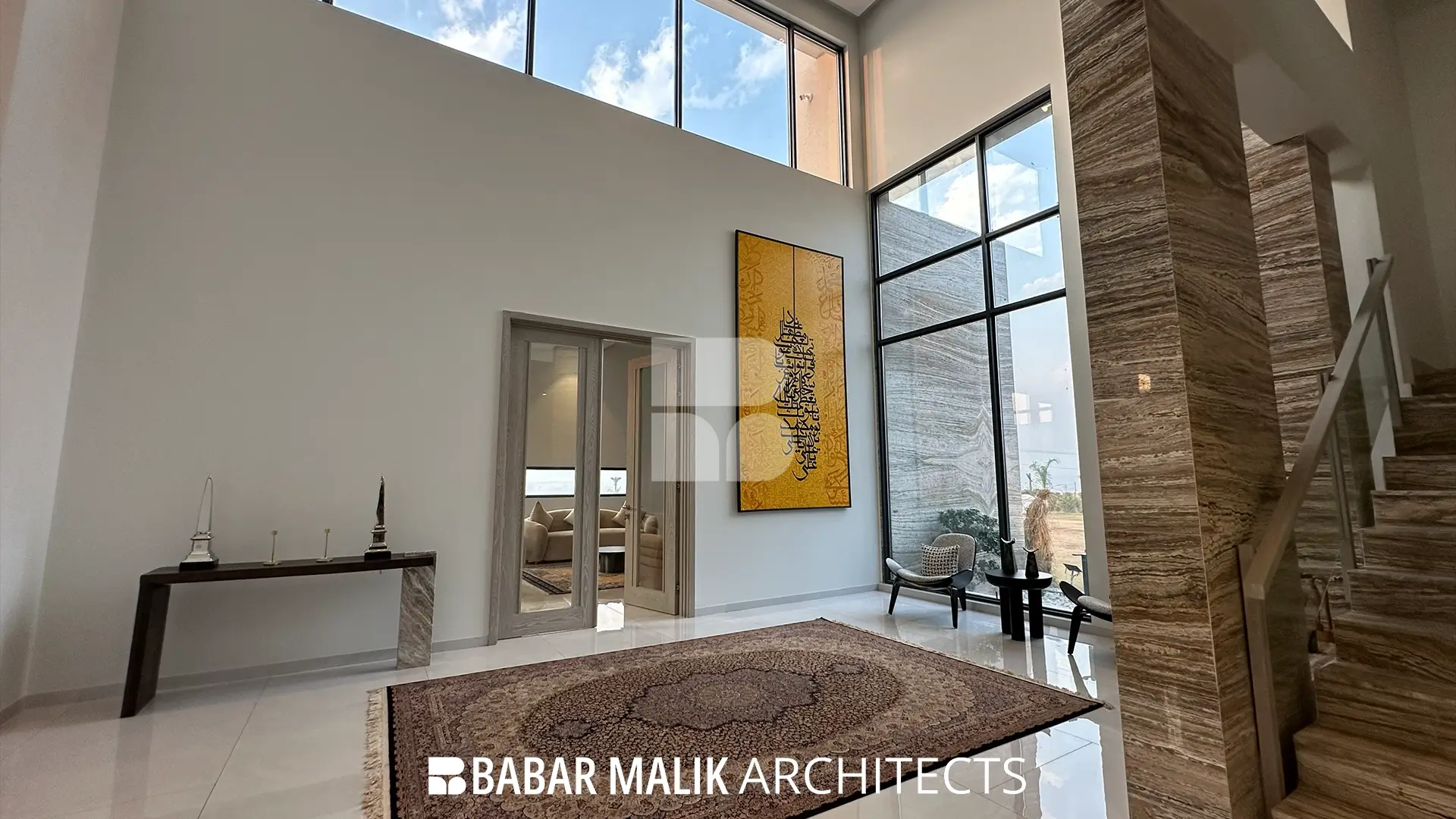 A grand double-height foyer with large floor-to-ceiling windows, featuring a structural column clad in travertine and a large-scale Islamic calligraphy artwork on the main wall.