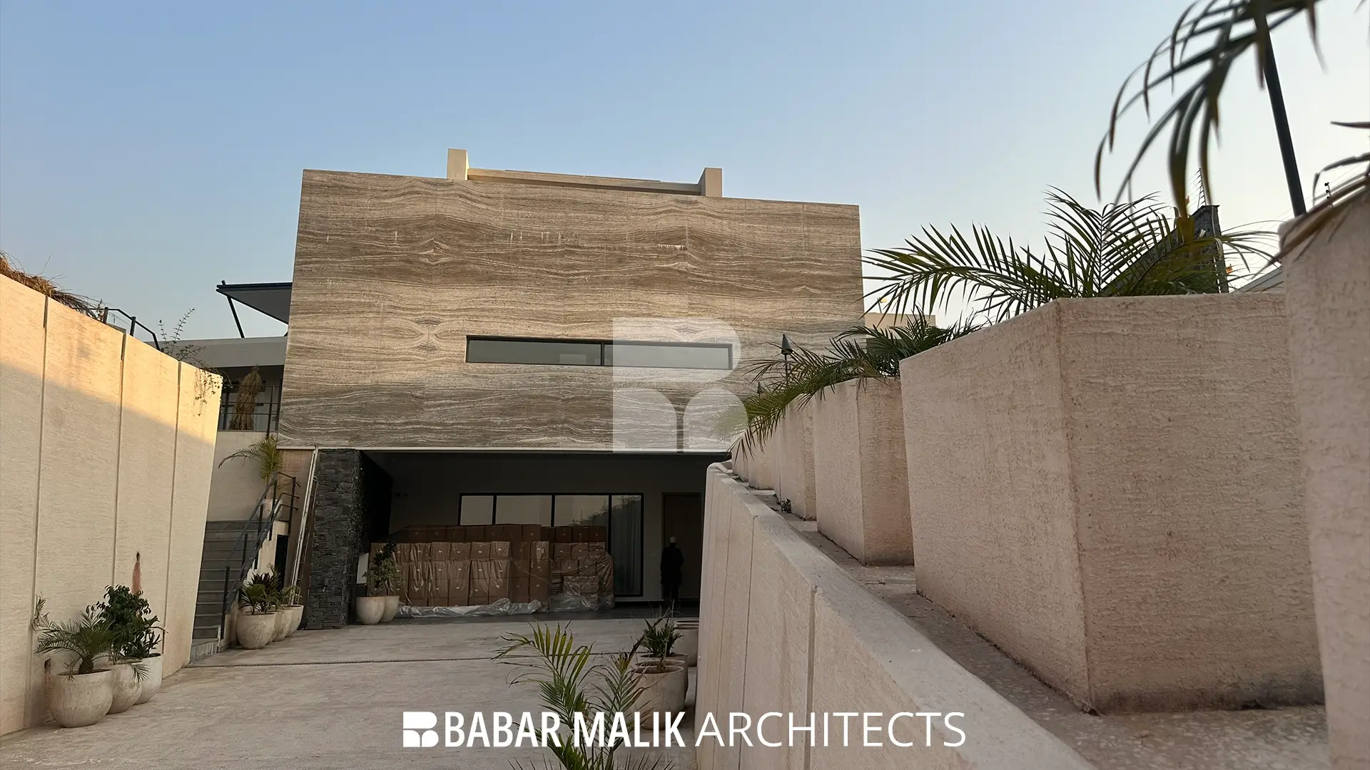 View of the descending driveway highlighting textured retaining walls and stepped planters leading towards the covered garage entrance beneath the main travertine volume.