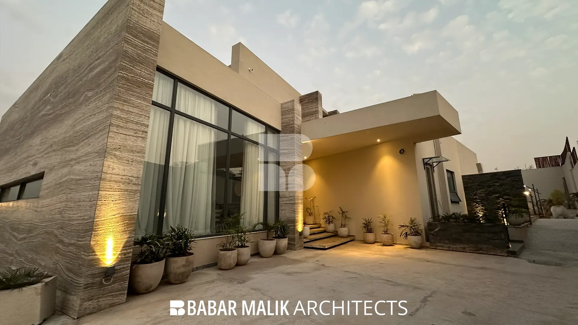 Ground-level exterior view of the house at dusk, showcasing large floor-to-ceiling windows, a covered entrance porch, illuminated planters, and travertine architectural columns.