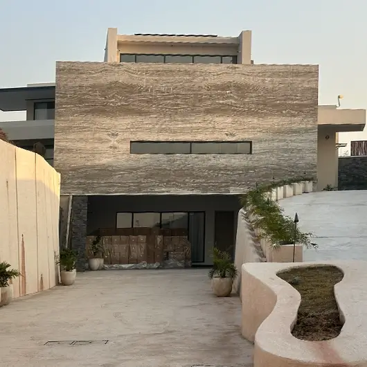 Front view of the residence showing the travertine facade, a descending concrete driveway leading to a lower-level garage, and an organically shaped built-in planter.
