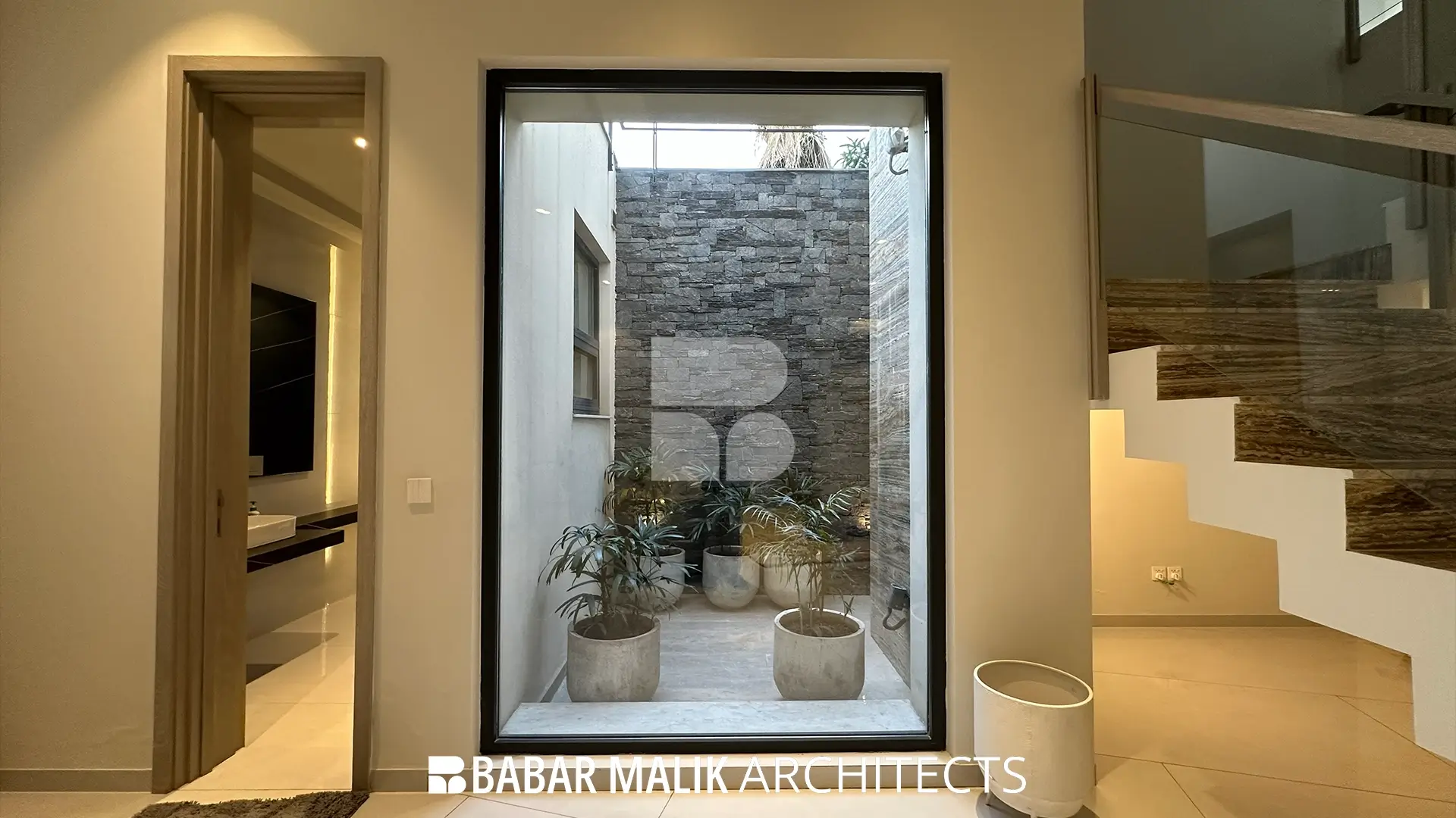 View from an interior hallway looking through a large, black-framed picture window into a central courtyard with dark stone feature walls and potted plants.