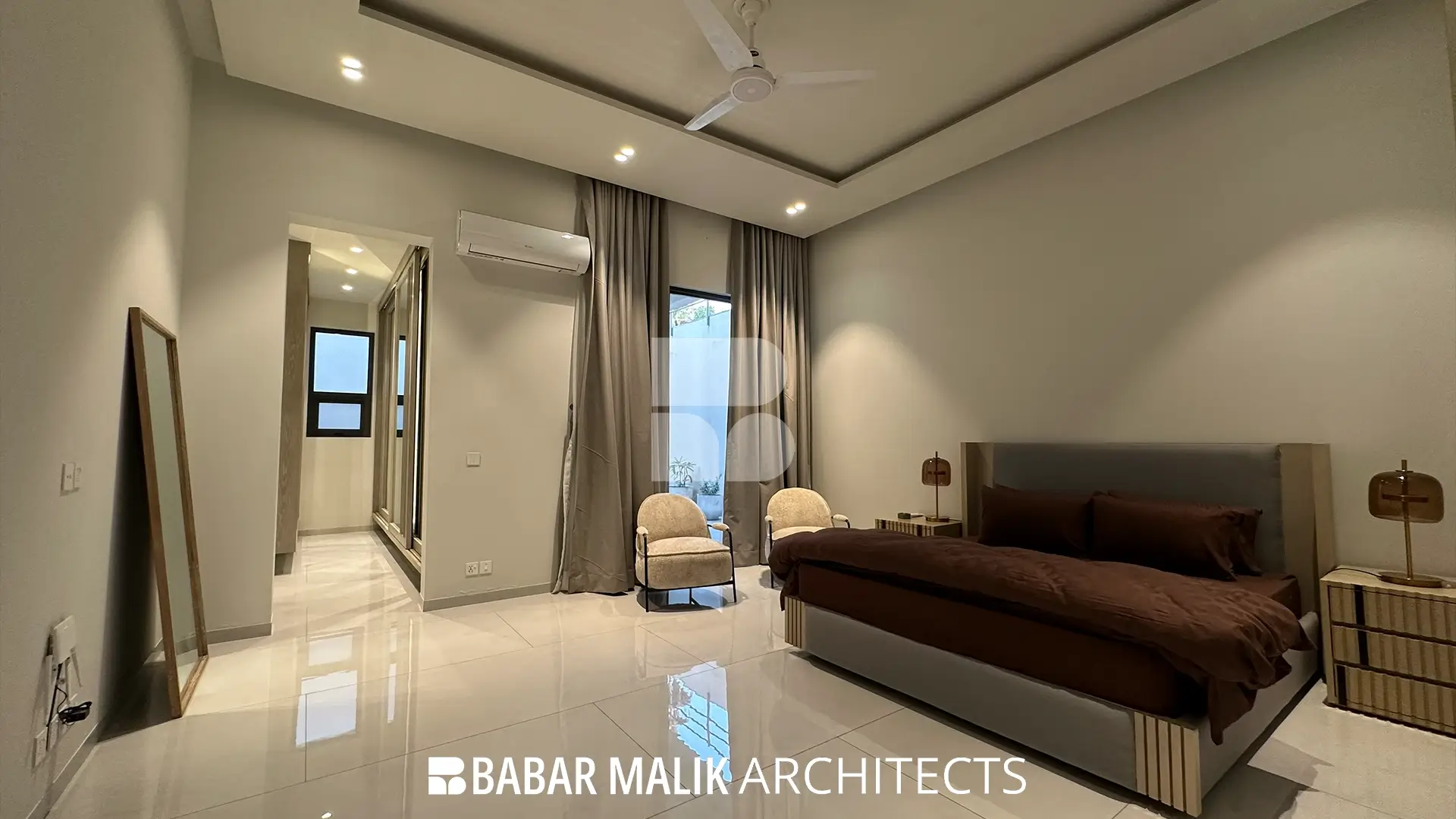 Master bedroom interior with light neutral floor tiles and walls, featuring a bed with brown linens, two textured accent chairs, and full-height curtains over a glass door.