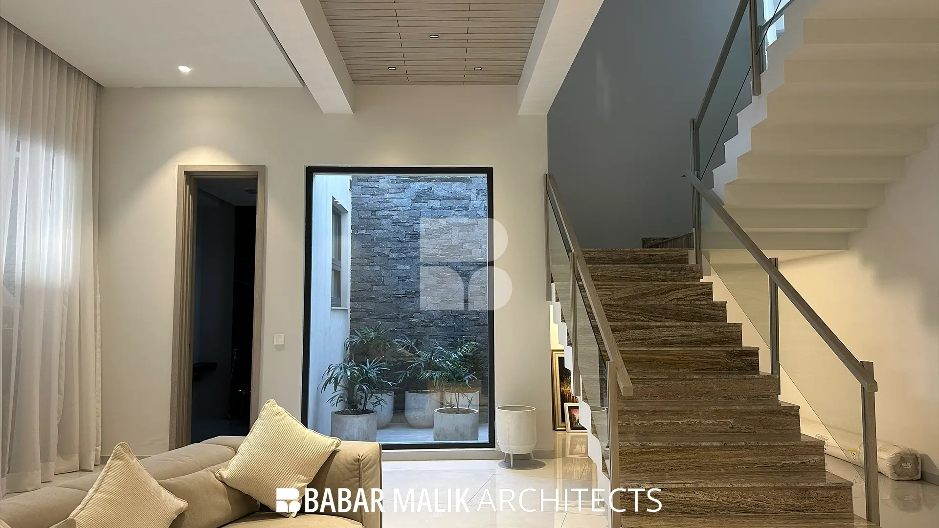 Open-plan living space positioned next to a stone staircase, featuring neutral beige seating, a wooden slatted ceiling element, and views into an enclosed stone courtyard.