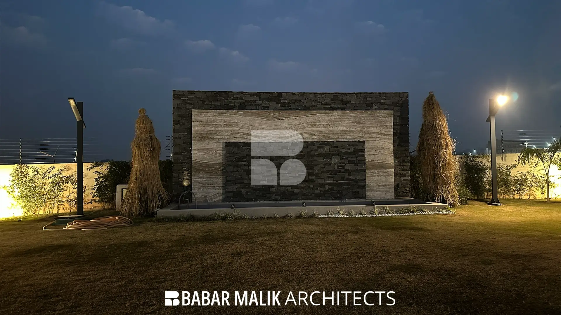 A standalone outdoor water feature illuminated at night, constructed with a combination of dark, rough-hewn stone and smooth travertine bands, flanked by landscaping.