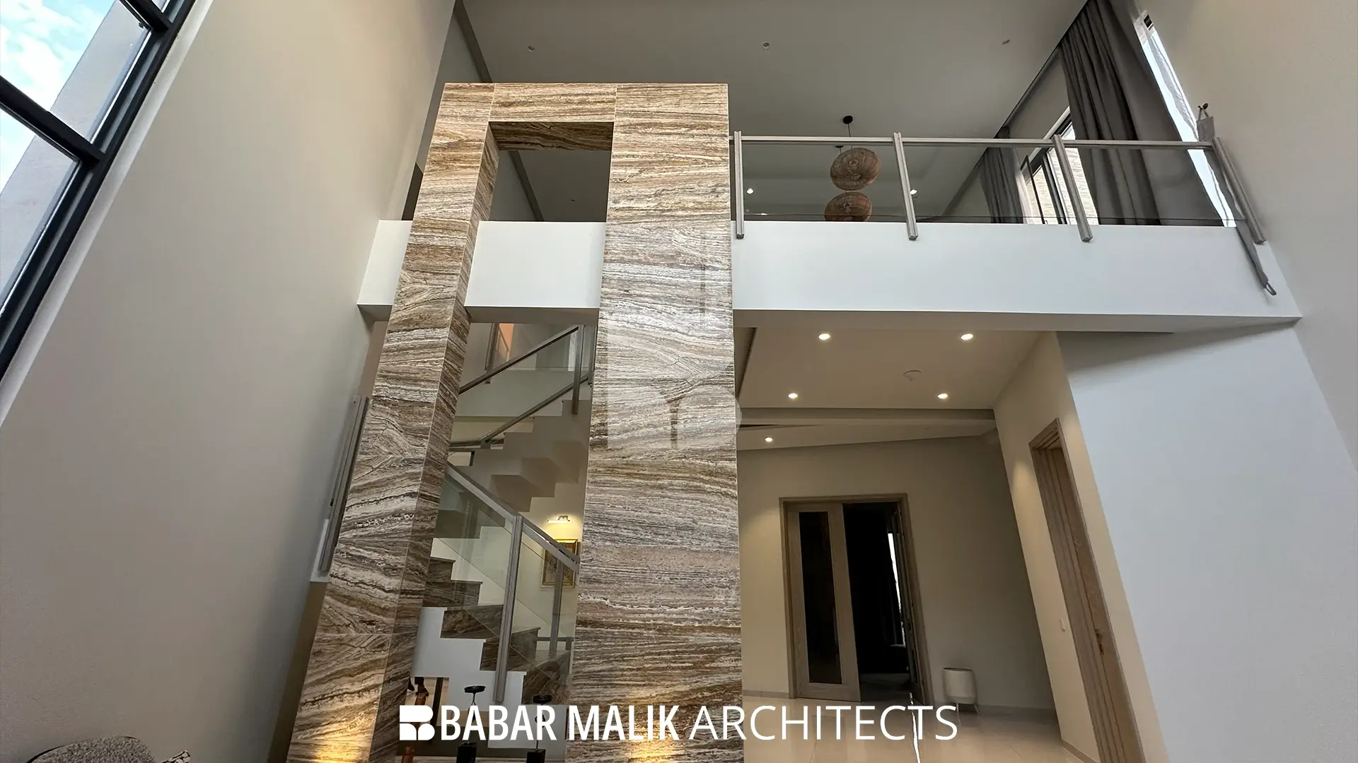 A double-height interior space dominated by a prominent architectural feature clad in travertine, integrating with the staircase and upper-level gallery with glass railings.
