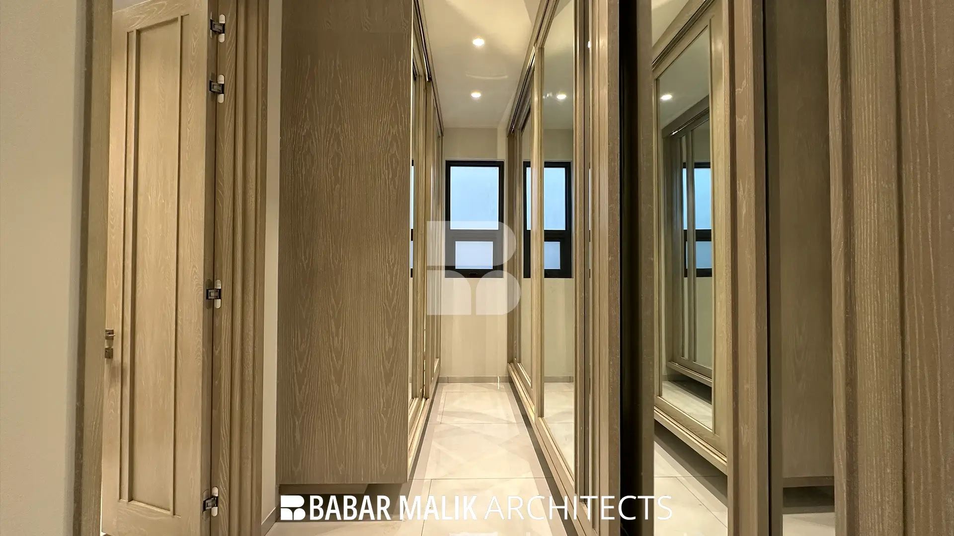 A narrow walk-in closet corridor flanked by floor-to-ceiling wooden wardrobes, featuring mirrored doors on one side to visually expand the space.